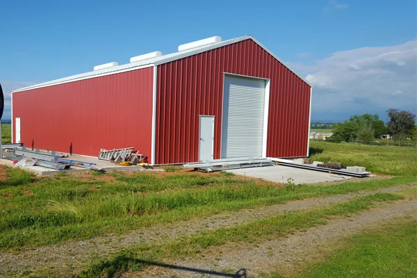 Red metal agricultural building with a large roll-up door, surrounded by green grass and gravel pathway, showcasing Ibarra Construction's metal building services in Woodland, CA.
