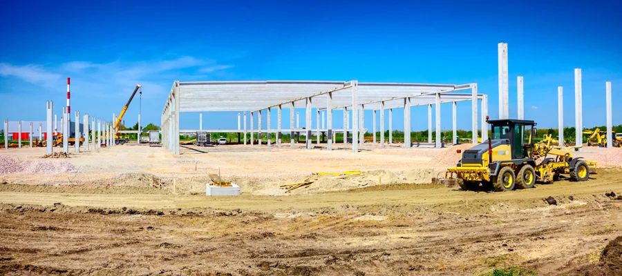 Construction site with heavy machinery grading land, steel framework for a building under construction, clear blue sky, and surrounding earthwork for optimal drainage and foundation stability.