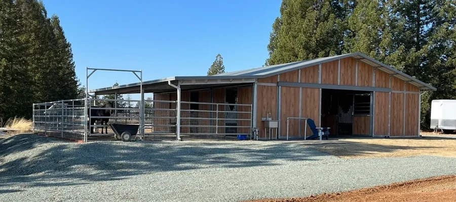 Metal building structure with wooden siding, designed for equestrian use, featuring a fenced area, gravel foundation, and surrounded by trees, illustrating Ibarra Construction's metal building installation services in Woodland, CA.