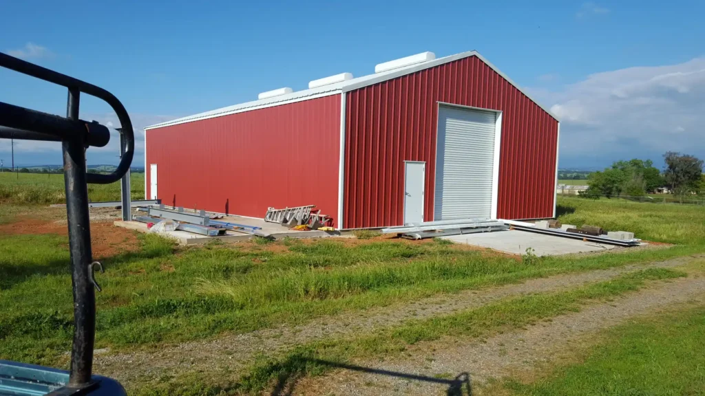 Red metal barn structure under construction with a large roll-up door and foundation work visible, set against a grassy landscape, showcasing Ibarra Construction's expertise in custom barn installation.