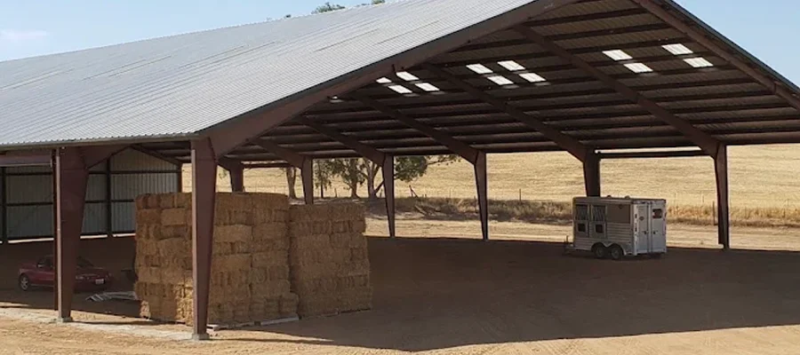 Steel structure barn with open design, featuring stacked hay bales and a horse trailer inside, showcasing Ibarra Construction's expertise in durable agricultural buildings.