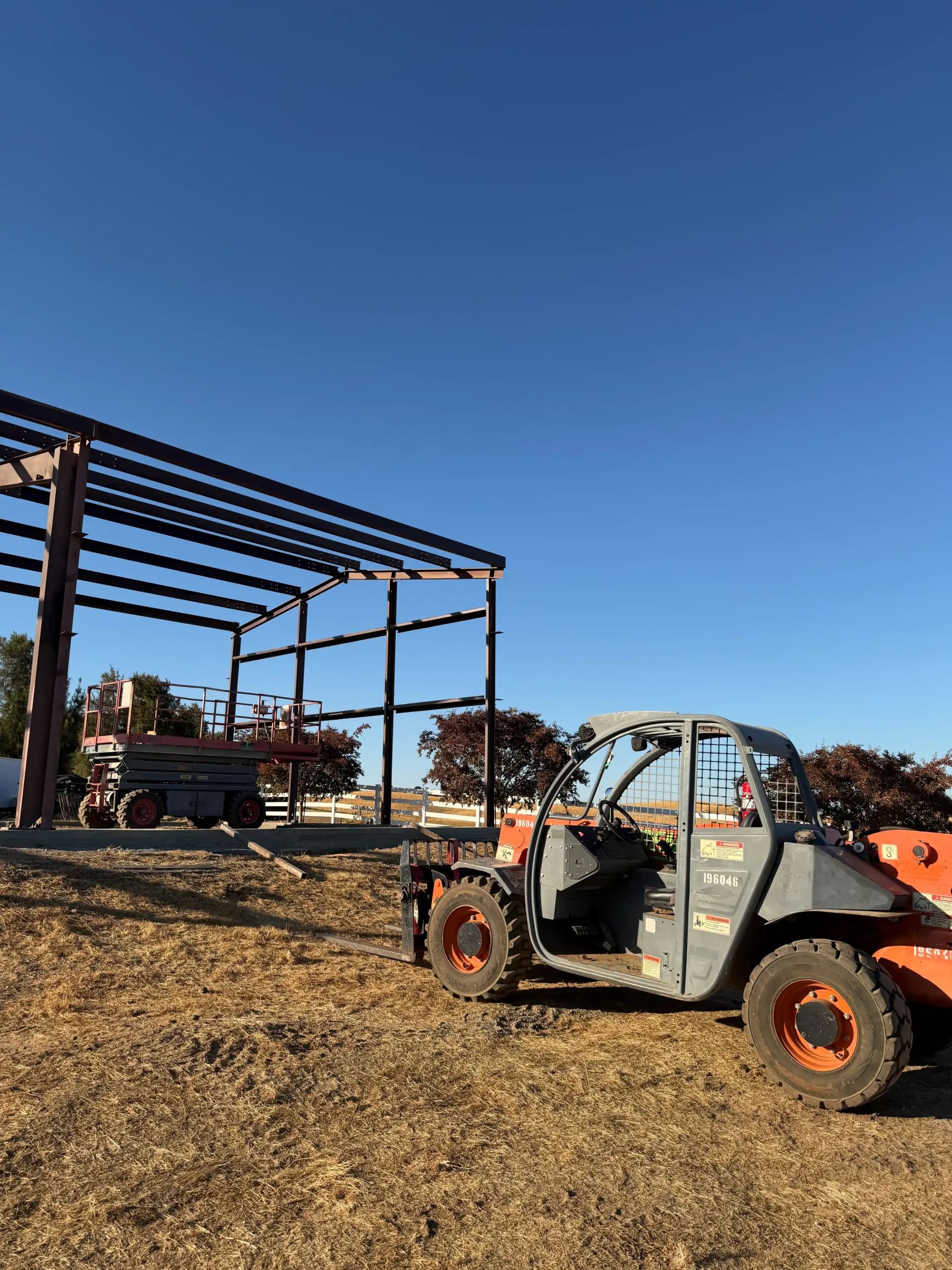 Construction site in Vacaville, CA, featuring a partially constructed residential steel building with a metallic frame, heavy machinery, and a clear blue sky.