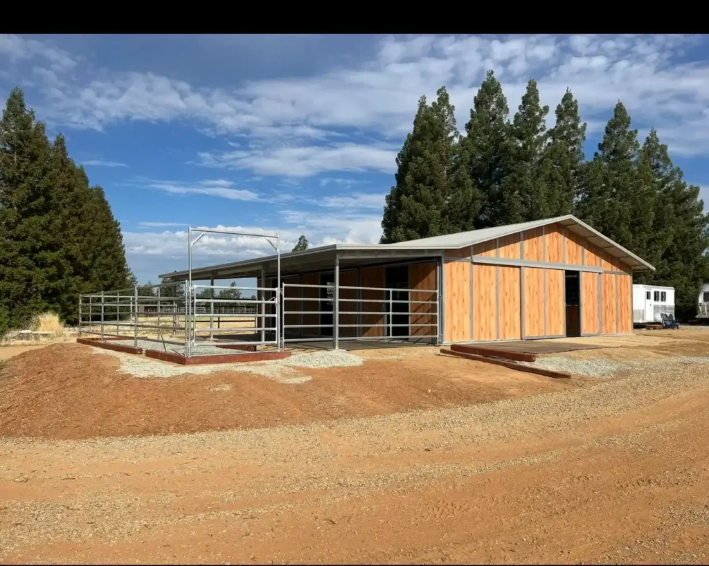 Residential horse barn in Wilton, CA, featuring a steel structure, wooden siding, and a concrete foundation, surrounded by natural landscaping and clear blue skies.