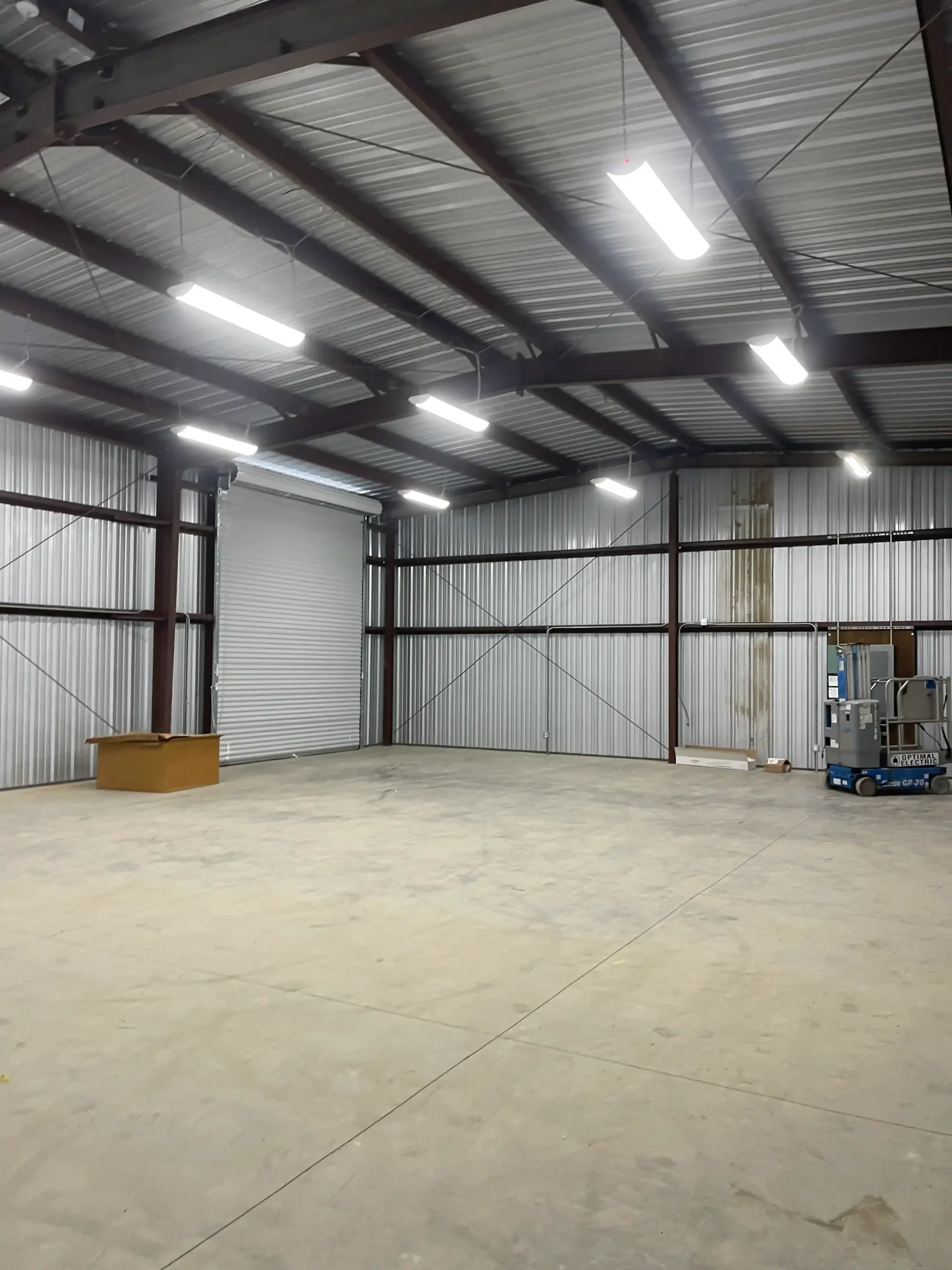 Interior of a commercial metal building featuring exposed steel beams, bright overhead lighting, and a spacious floor area with a storage box and equipment in the corner, illustrating flexibility and durability for business applications.