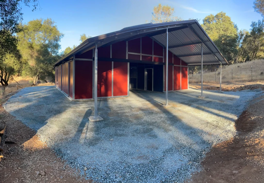Residential horse barn with a steel structure and custom concrete foundation, surrounded by gravel and trees in Plymouth, CA, showcasing Ibarra Construction's craftsmanship.