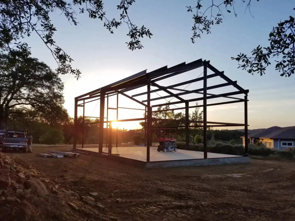 Metal building framework under construction at sunset, showcasing site preparation and foundation work, with equipment and vehicles nearby.